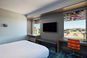 Habitación de hotel con cama, escritorio, silla, televisión y ventana con vista a la ciudad.