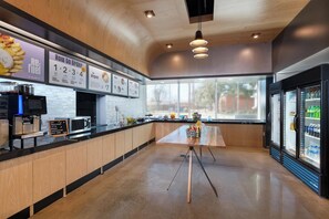 A modern kitchen area with a long counter, a coffee machine, a microwave, and a refrigerator. There are menu boards above the counter and a window providing natural light. - Aloft by Marriott Waco Downtown (Waco)