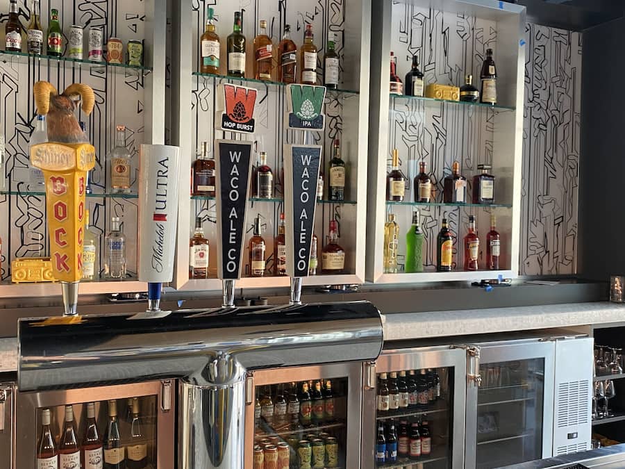 Zona de bar con múltiples grifos etiquetados con diferentes tipos de alcohol, estantes llenos de botellas y un letrero de neón que dice 'WXYZ'.
