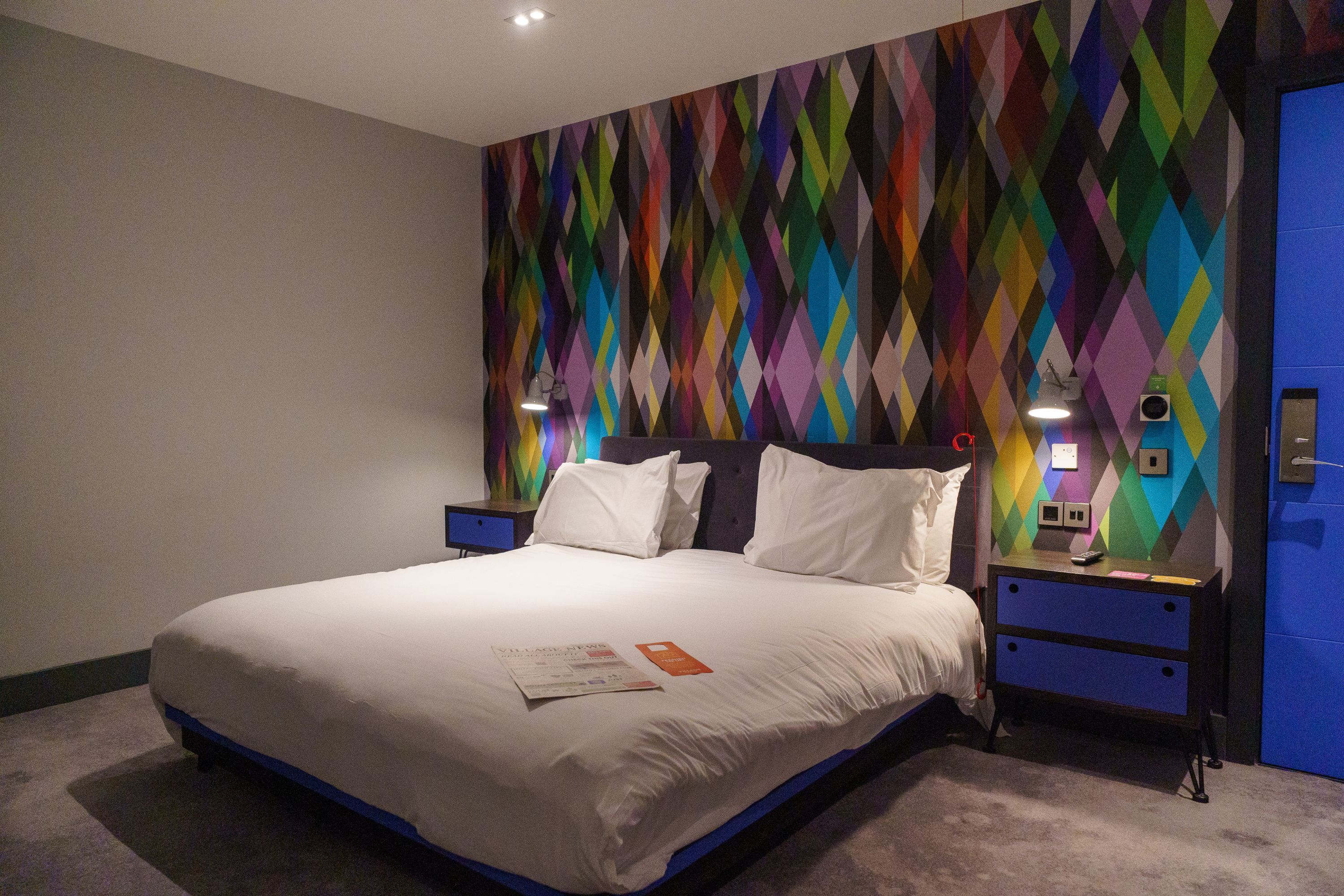 Una habitación de hotel con una cama grande, una mesita de noche, una lámpara de pared y una puerta con un panel azul.