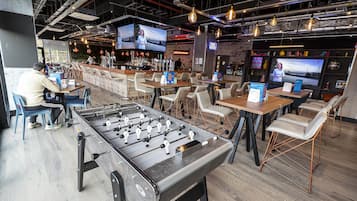 Un espace bar moderne offrant de nombreuses options d’assise, une table de baby-foot et de grands écrans diffusant du contenu.