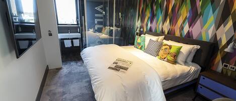 A modern bedroom with a large bed, a glass door leading to a balcony, and a colorful geometric wall pattern.