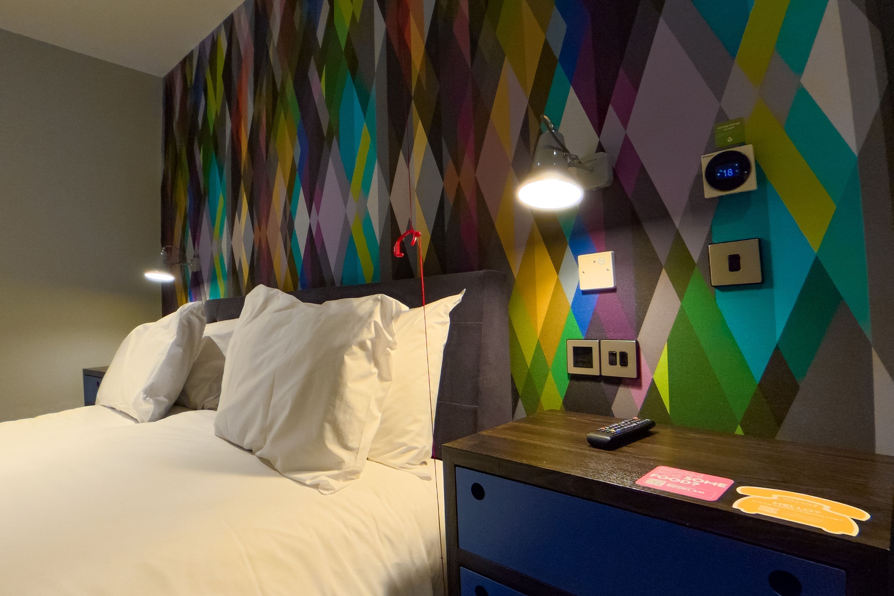 Une chambre d’hôtel avec un lit, une table de chevet avec une télécommande, une horloge murale et une prise électrique.