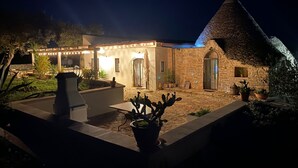 Ein Steinhaus mit einer gefliesten Terrasse, einer überdachten Sitzgelegenheit und Kübelpflanzen.
