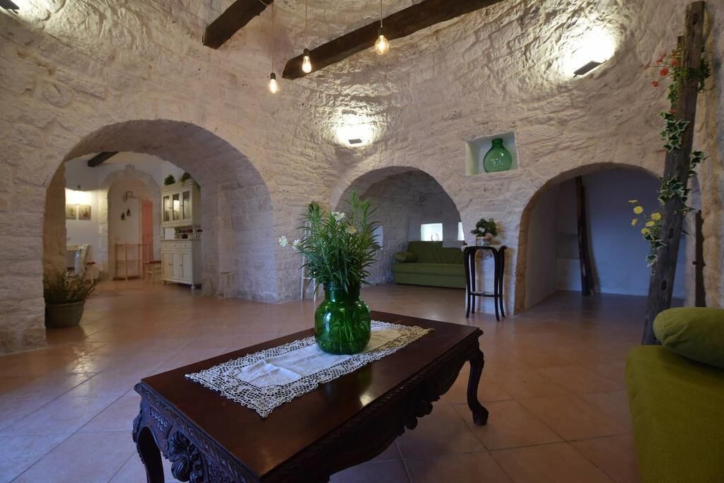 Un intérieur rustique avec des murs en pierre, une table en bois recouverte d’une nappe en dentelle et un vase vert orné de fleurs.