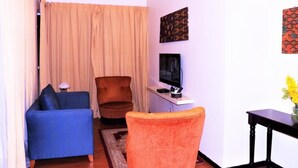 A living room with a blue sofa, an orange armchair, a TV on a stand, and a small table with a vase of flowers. - Maytower Apartment (Kuala Lumpur)
