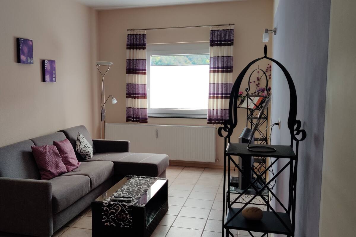 A living room with a sofa, a coffee table, and a decorative stand. There is a window with curtains and a radiator on the wall.