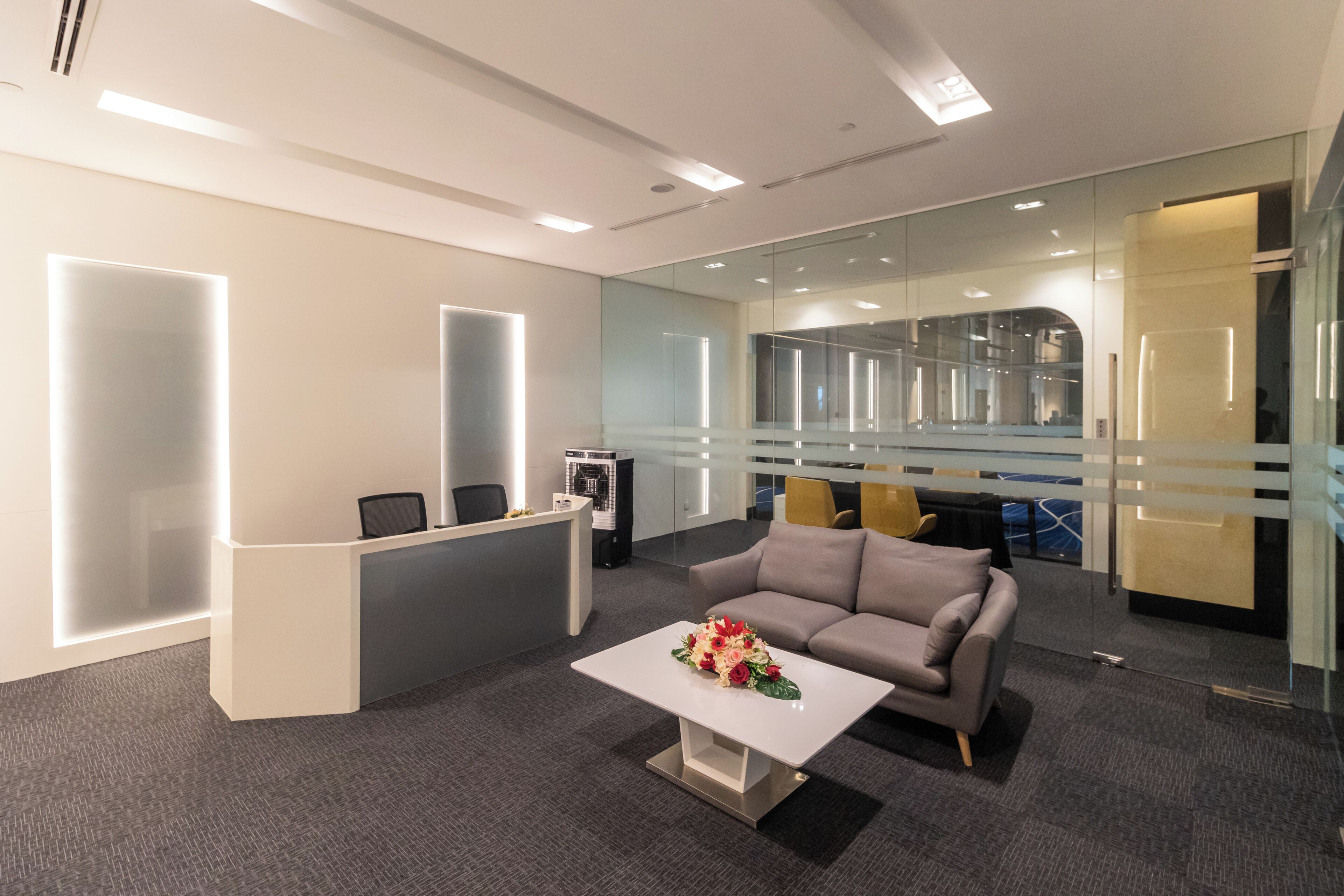 A modern office reception area with a reception desk, a grey sofa, and a coffee table with flowers.