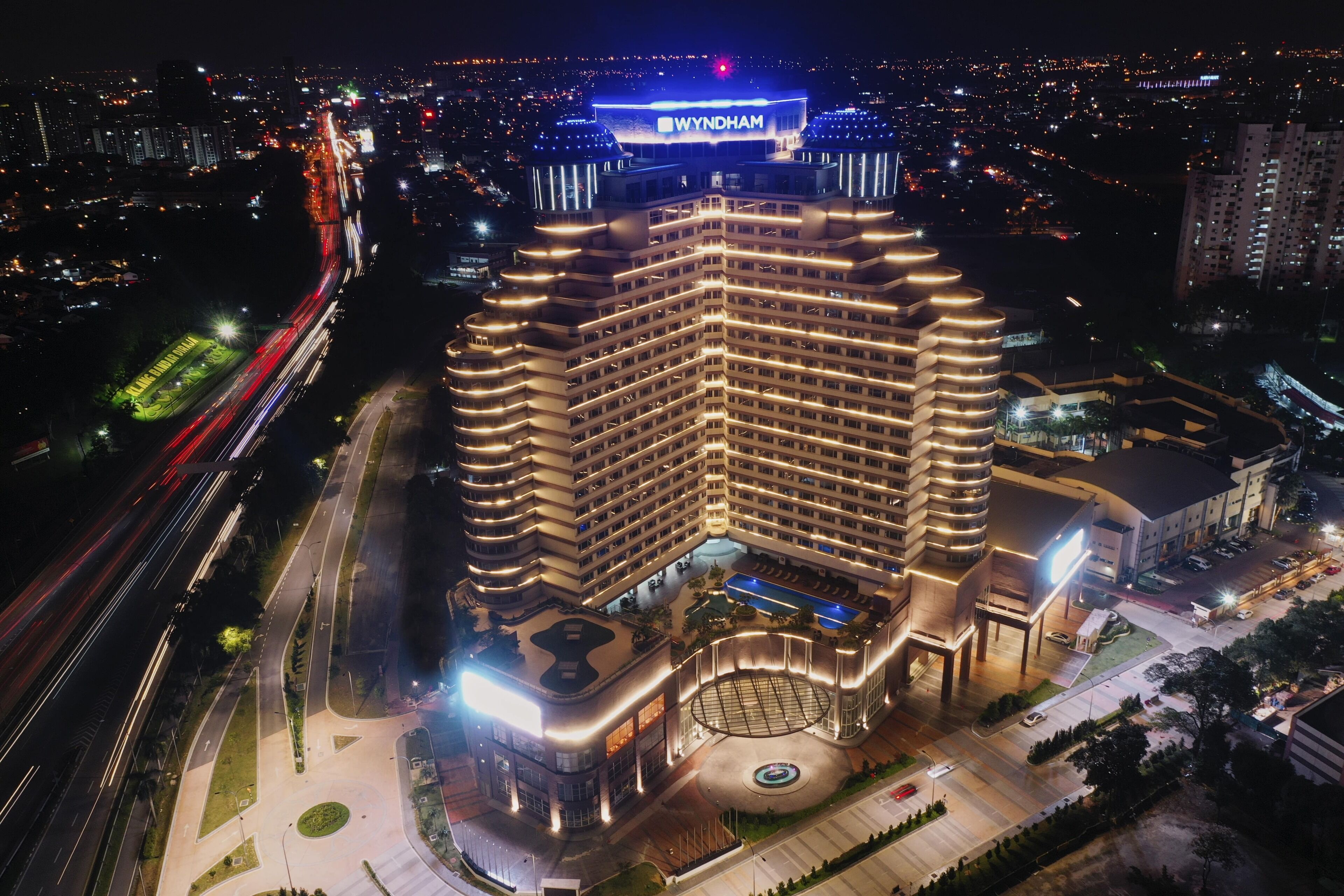 Een meerhoog hotel met een opvallend 'WYNDHAM' bord, omgeven door verlichte wegen en een aangelegde omgeving met een ronde tuin.