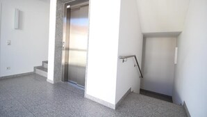 Um elevador moderno com acabamento metálico, um pequeno conjunto de escadas levando a um patamar, e um corrimão no lado direito.