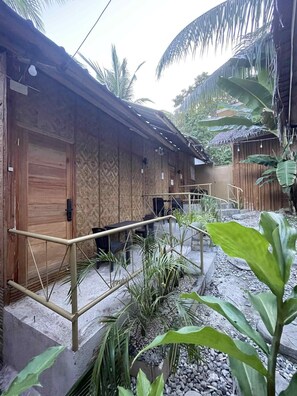 Un bâtiment en bois, doté d’un toit de chaume, entouré de plantes tropicales et bordé d’un chemin de gravier.
