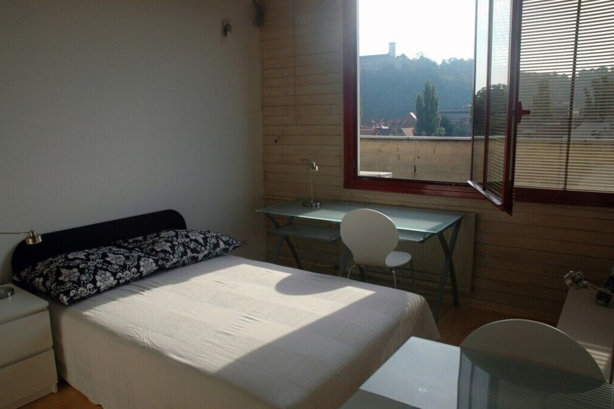 Une chambre à coucher avec un lit, un bureau et une chaise. Il y a une fenêtre avec des stores et, de la fenêtre, on peut voir des bâtiments et des arbres.