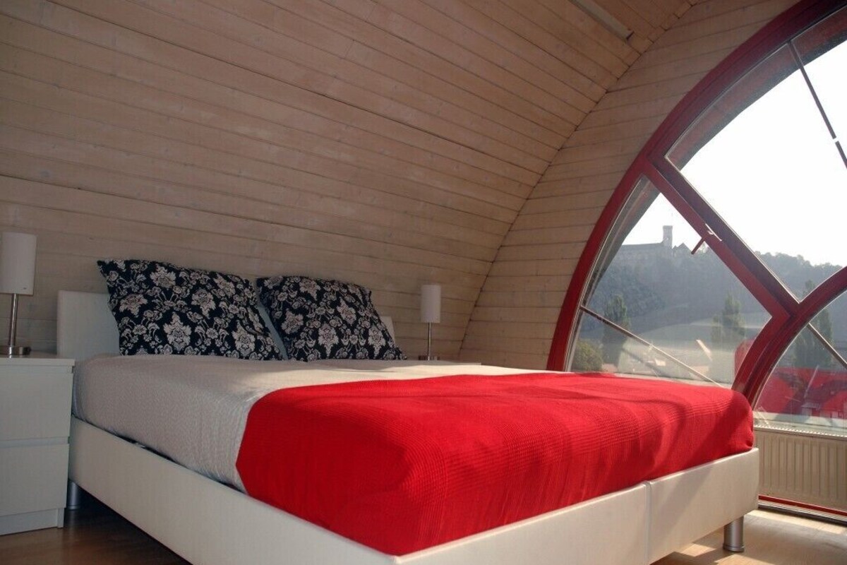 Une chambre avec un plafond en pente, un lit recouvert d’une couverture rouge, une table de chevet, une lampe et une fenêtre donnant sur un immeuble.