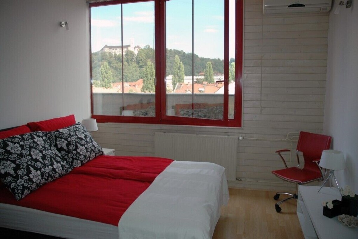 Une chambre avec une grande fenêtre, un lit avec une literie rouge et blanche, une chaise rouge et un radiateur.
