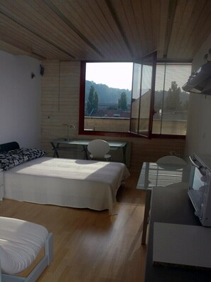 A bedroom with a bed, desk, chair, and a large window offering a view of the outdoors.