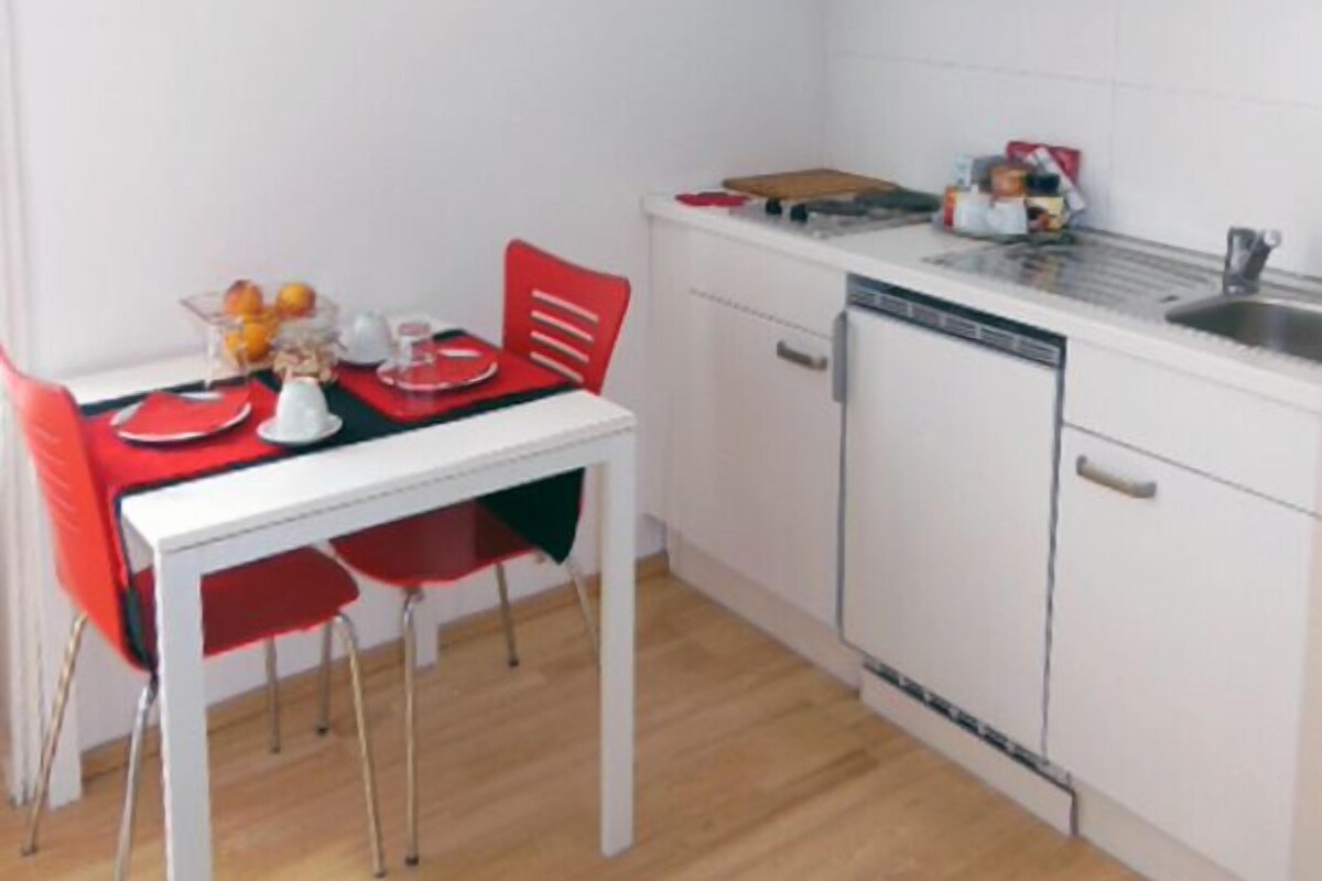 Une cuisine compacte avec des meubles blancs, un lave-vaisselle et un coin repas comprenant une chaise et une table rouges.