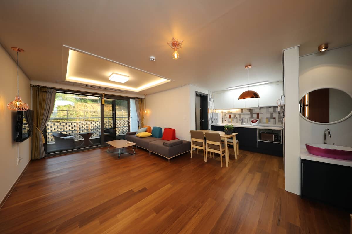 A modern living room with a sofa, chairs, and a dining area. It includes a kitchen with a sink and a large window.