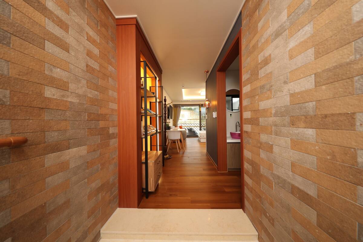 A narrow hallway with wooden flooring and brick walls, leading to a dining area with a table and chairs.