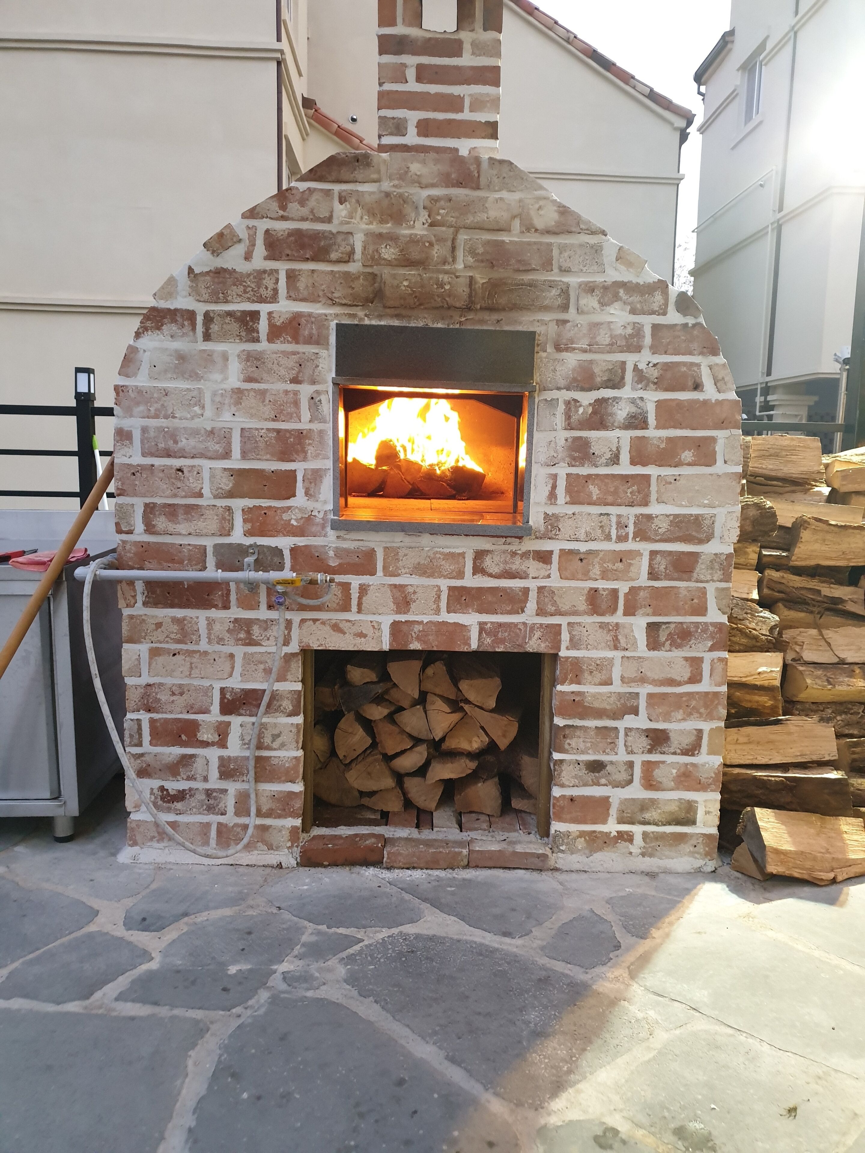 Ein aus Ziegeln gebauter Pizzaofen im Freien mit brennendem Feuer, umgeben von Steinböden und einem Holzstapel.