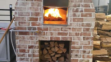 Un four à pizza extérieur en briques, avec un feu qui crépite à l’intérieur, entouré d’un sol en pierre et d’une pile de bois de chauffage.