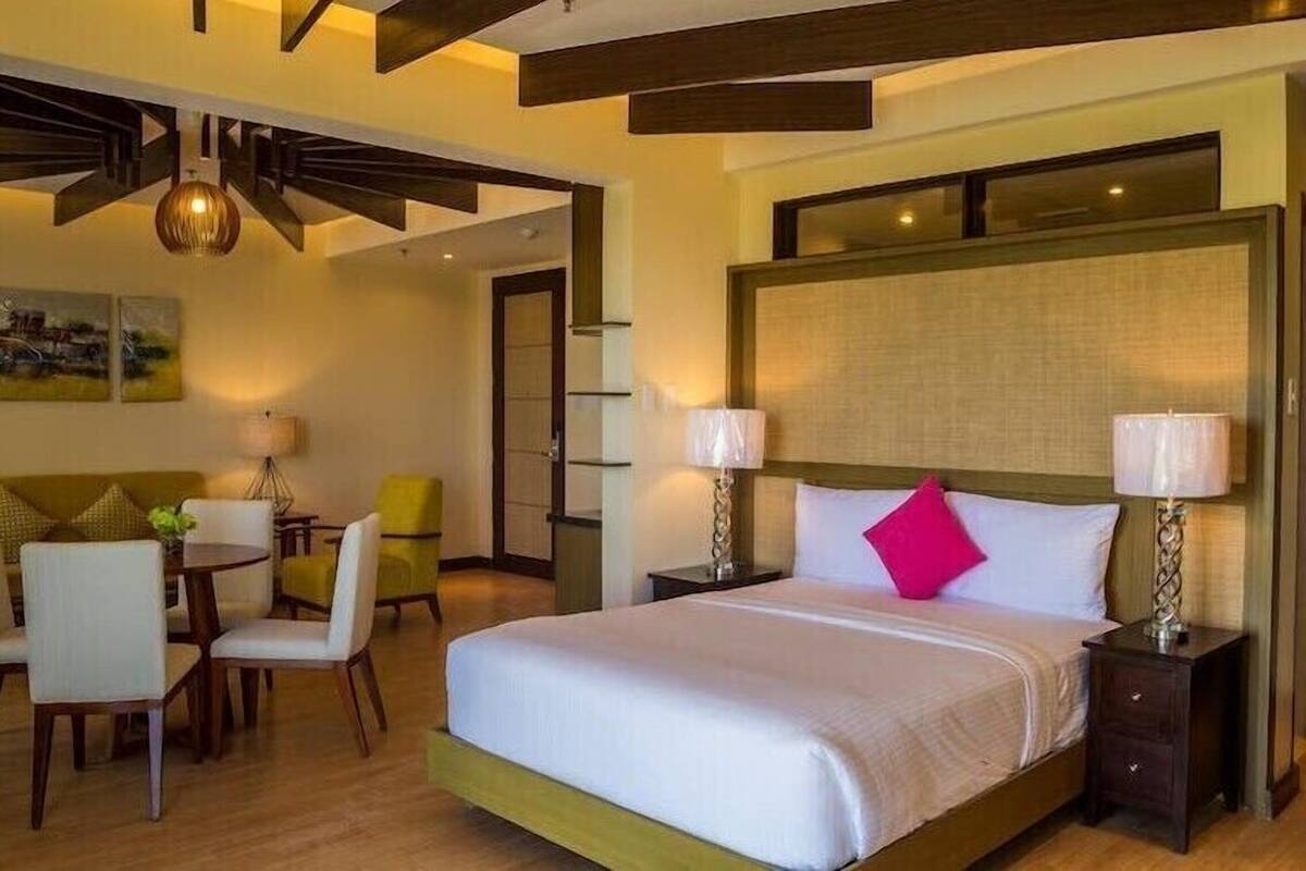 A hotel room with a large bed, two bedside tables, a desk with chairs, and a dining area.