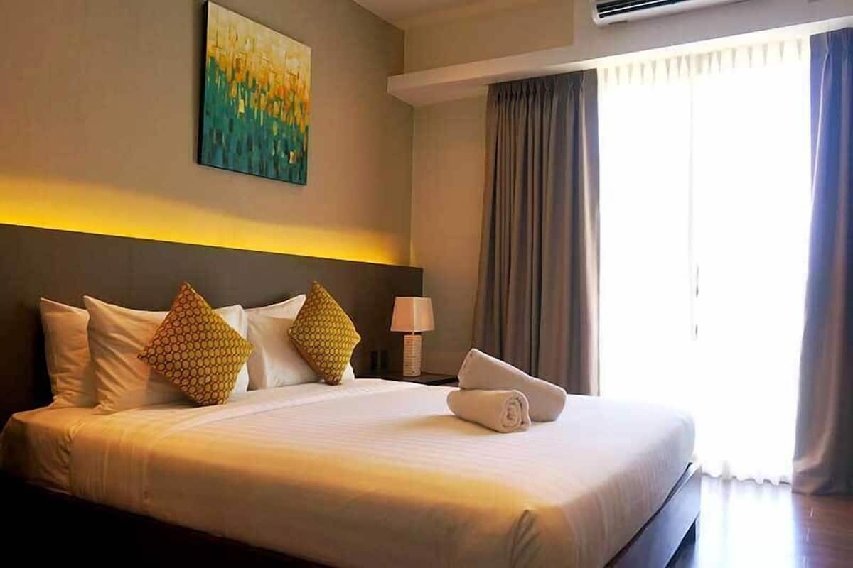 A hotel room with a large bed, a painting on the wall, a nightstand with a lamp, and a window with curtains.