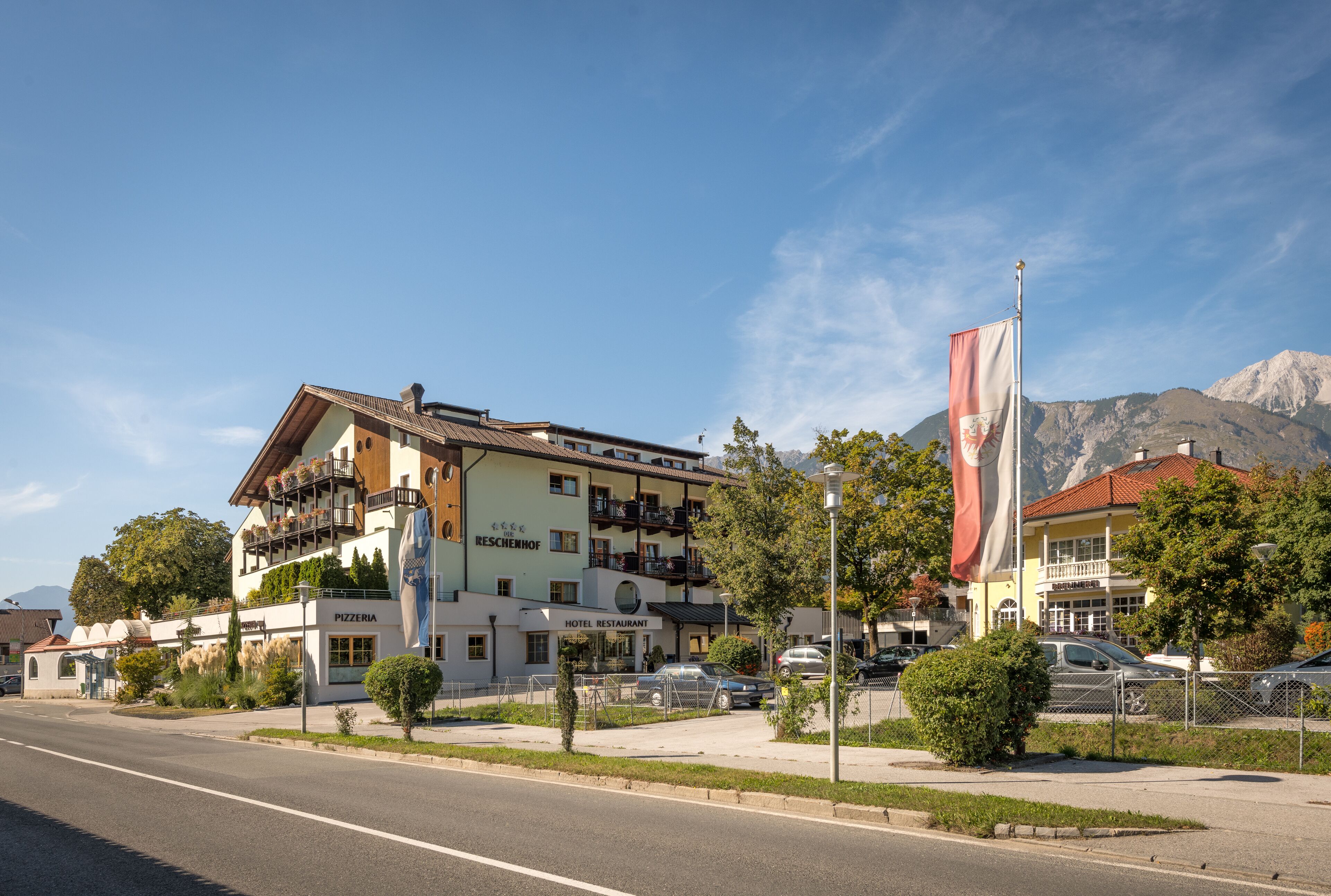 A hotel with a pizzeria and restaurant on the ground floor, surrounded by greenery and a mountain backdrop.