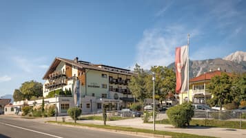 A hotel with a pizzeria and restaurant on the ground floor, surrounded by greenery and a mountain backdrop.