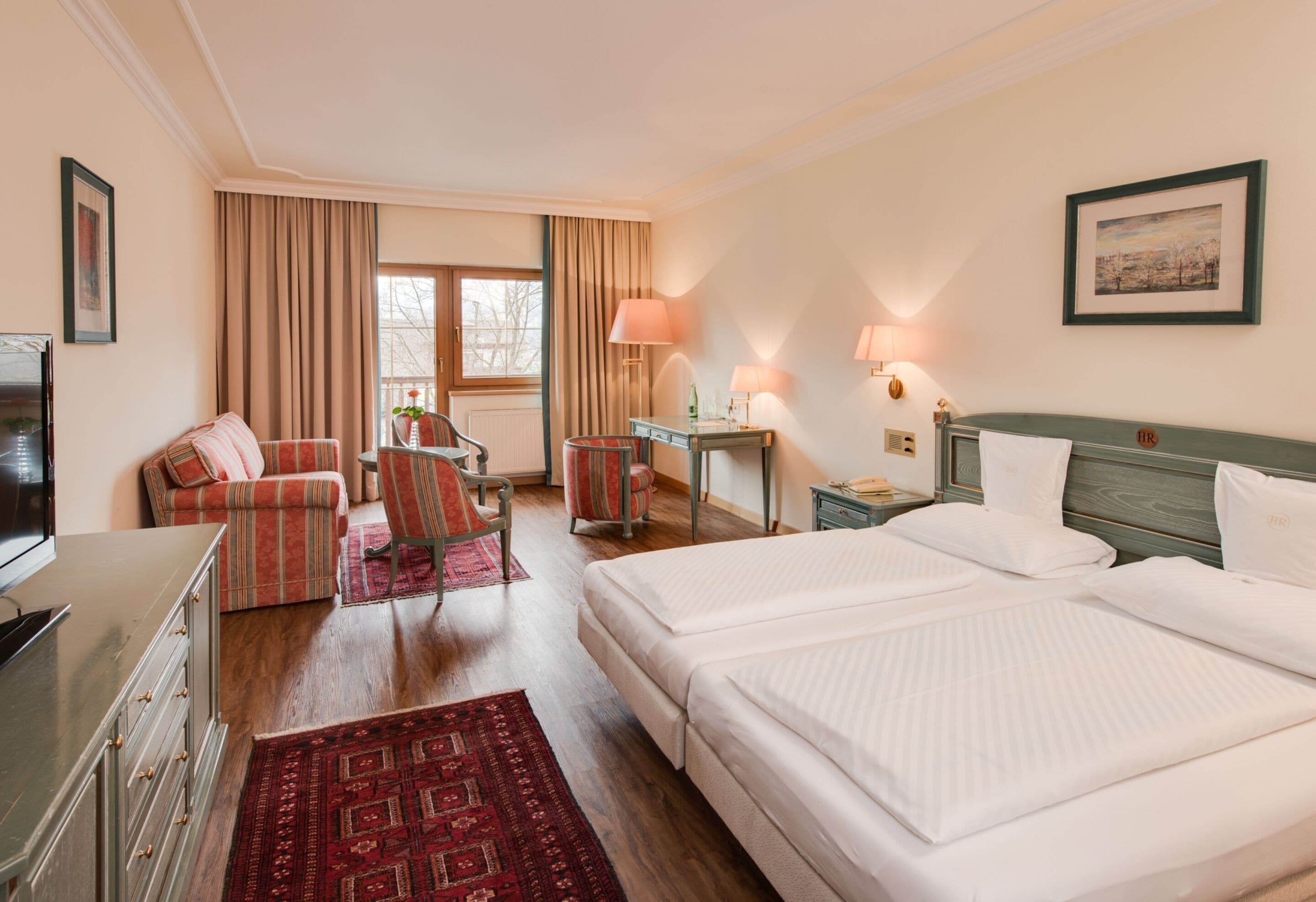 Quarto de hotel com cama, escrivaninha, cadeira, televisão e janela com cortinas.