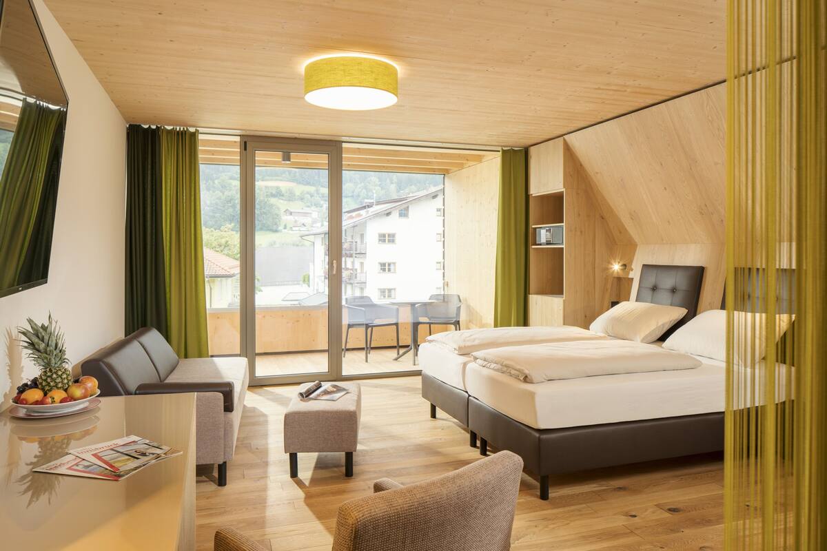 A modern hotel room with a bed, a desk with a chair, a small table with fruit, and a view of the outside.
