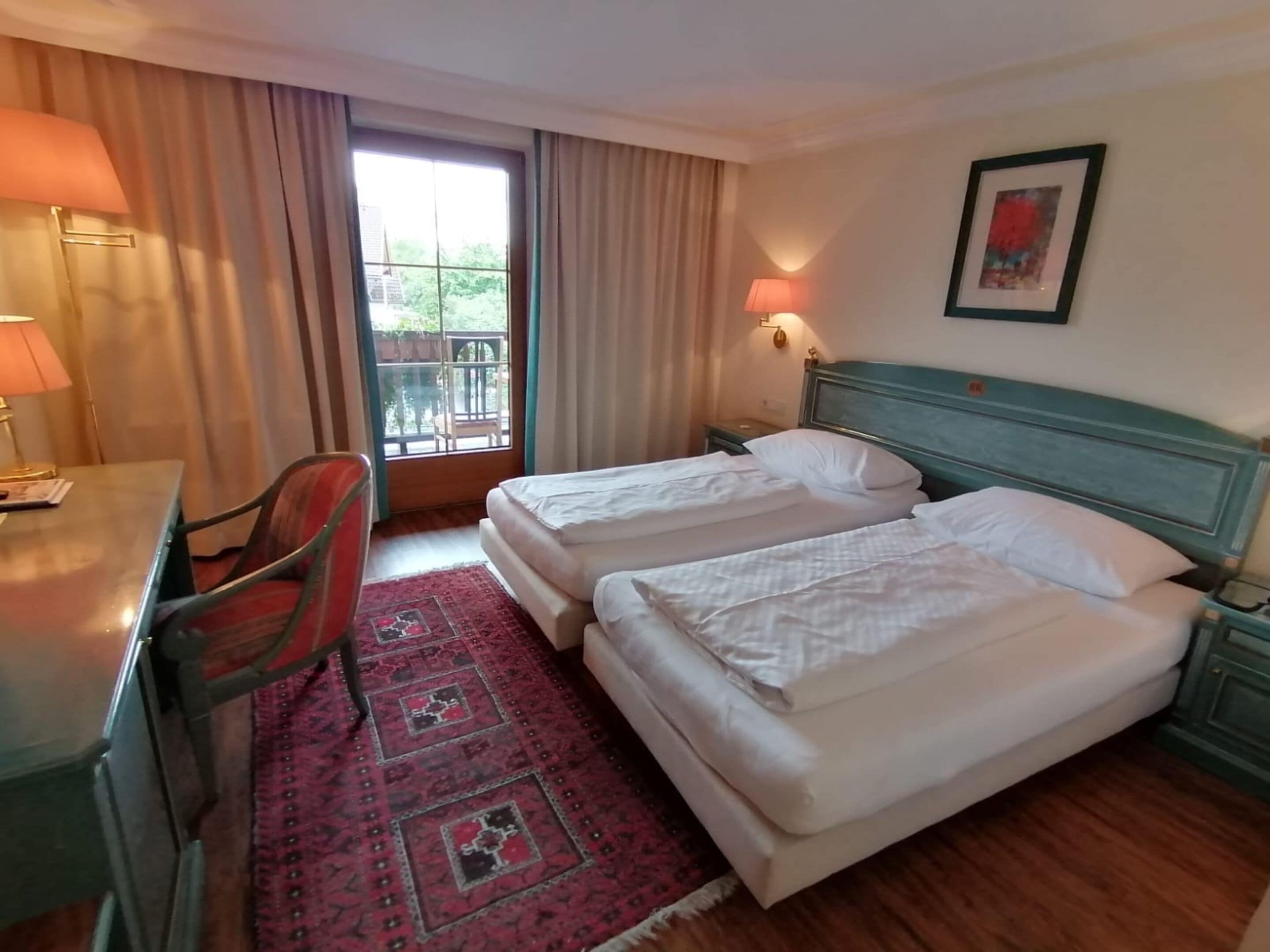 A hotel room with two beds, a desk, a chair, a window with curtains, a nightstand, and a wall-mounted lamp.
