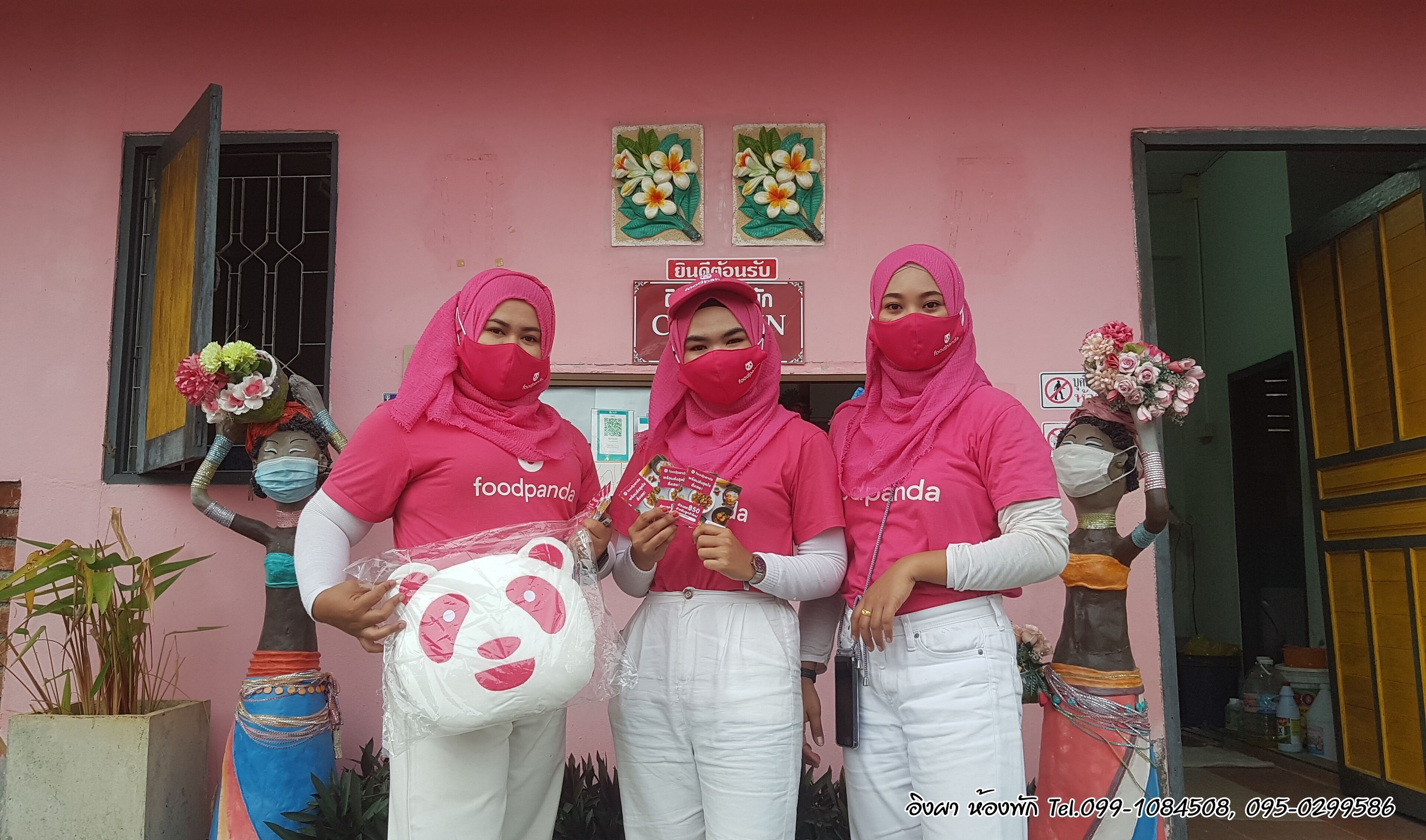 Three individuals in front of a building with a pink facade, wearing pink shirts and white pants, holding food items and flowers.