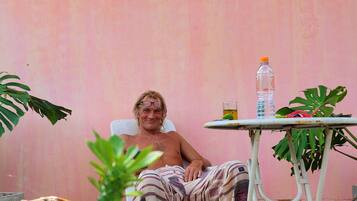A person seated on a chair in a room with a pink wall and a sign that reads "Angspha". There is a table with a bottle and a glass, and various plants around.