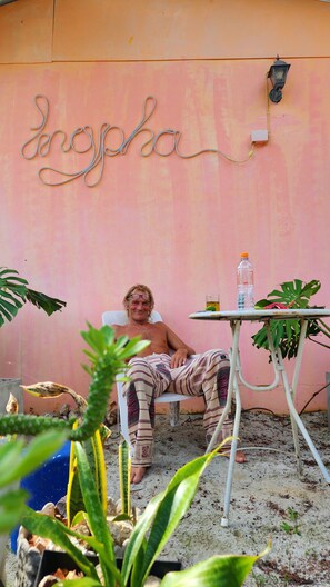 A person seated on a chair in a room with a pink wall and a sign that reads "Angspha". There is a table with a bottle and a glass, and various plants around.