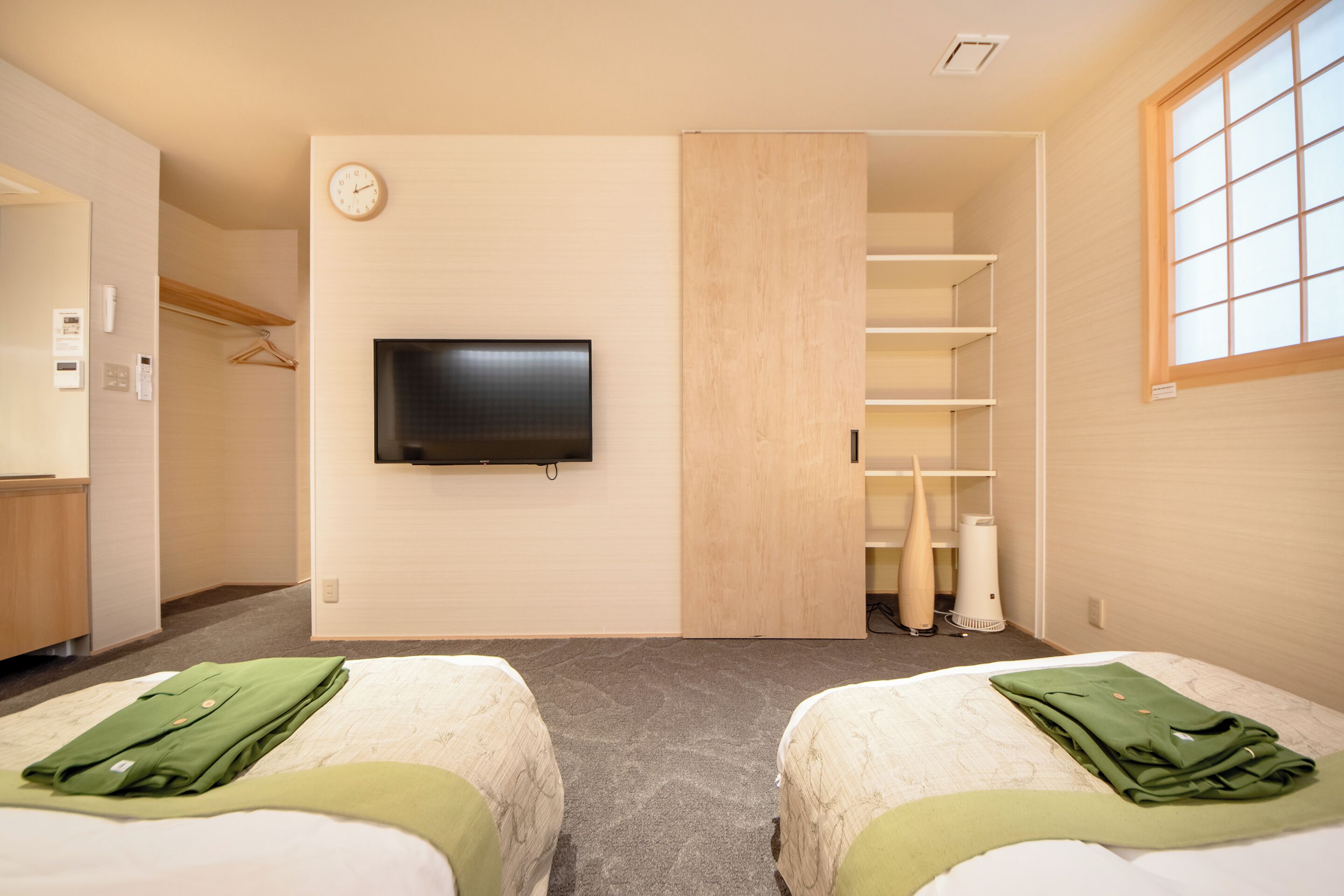 A hotel room with two beds, a TV mounted on the wall, a closet with shelves, and a window.