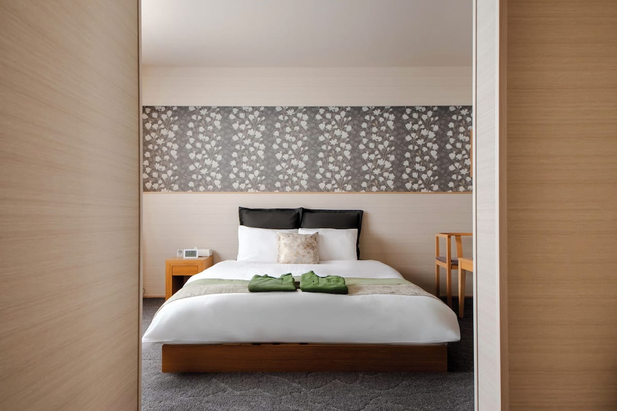 Una cama bien hecha con cabecera estampada con motivos florales, una mesita de noche con lámpara y una silla en una habitación de hotel moderna.