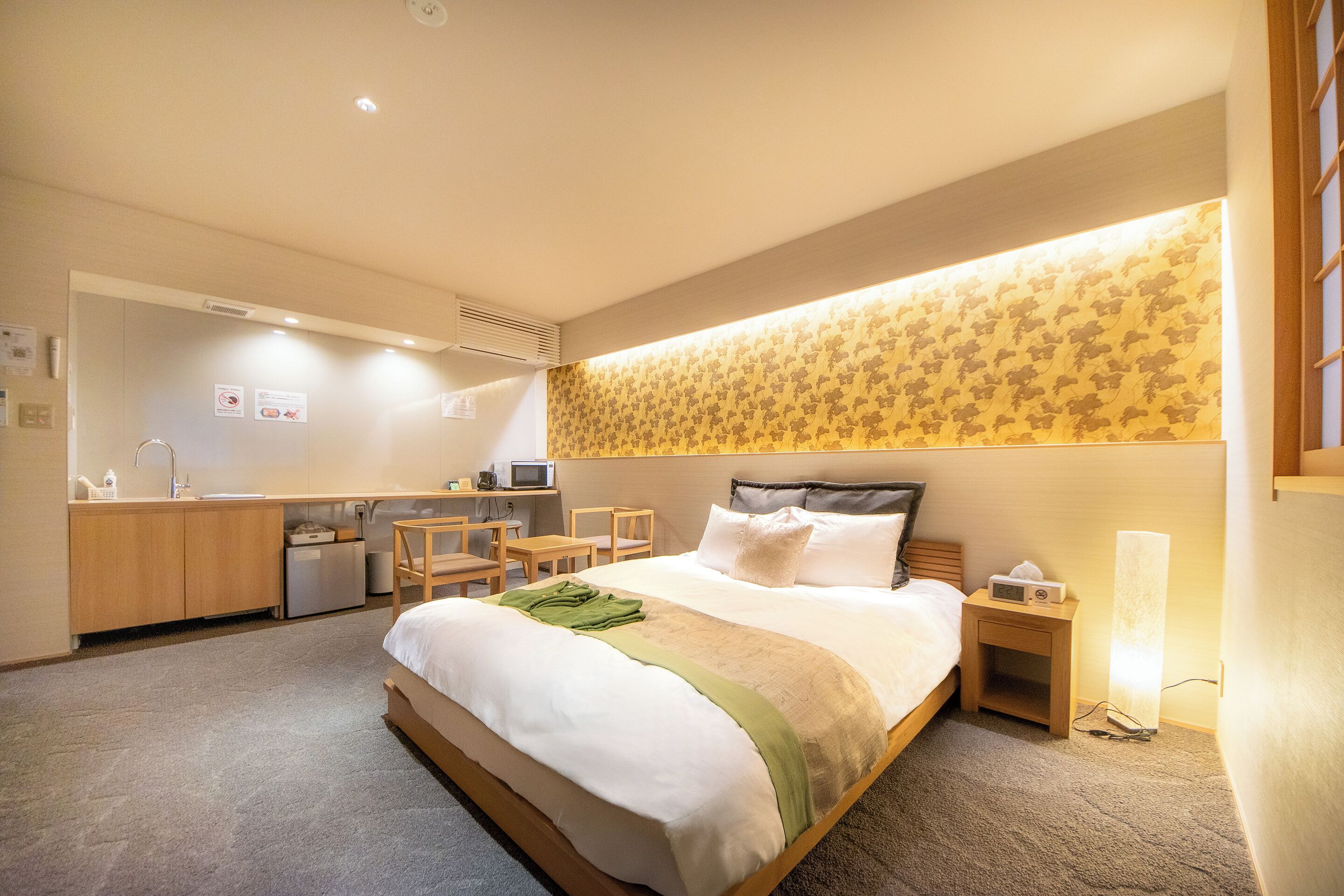 A hotel room with a bed, bedside table, lamp, and a small desk with a chair.