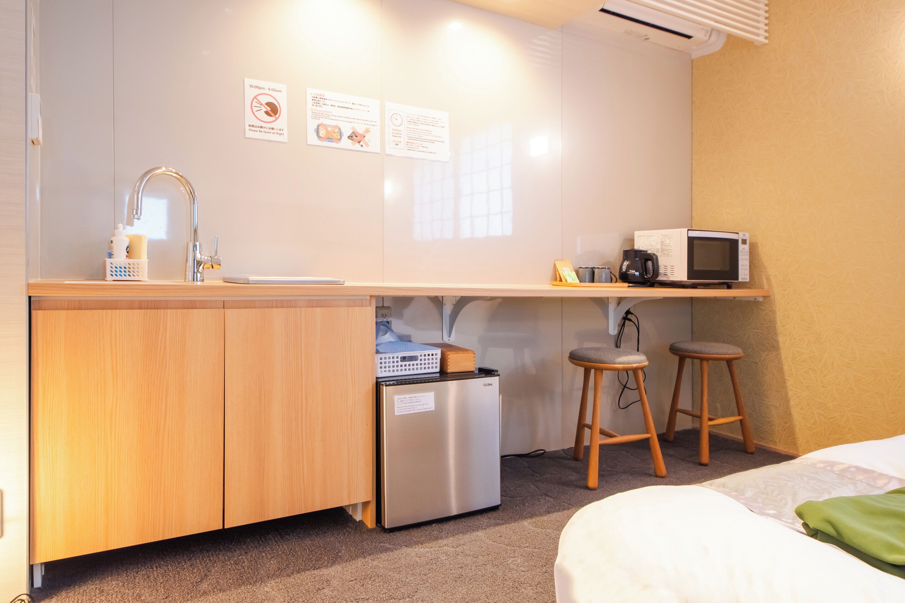 A compact kitchenette with a stainless steel refrigerator, a microwave, and a sink. There are two stools and a small table with a coffee maker.