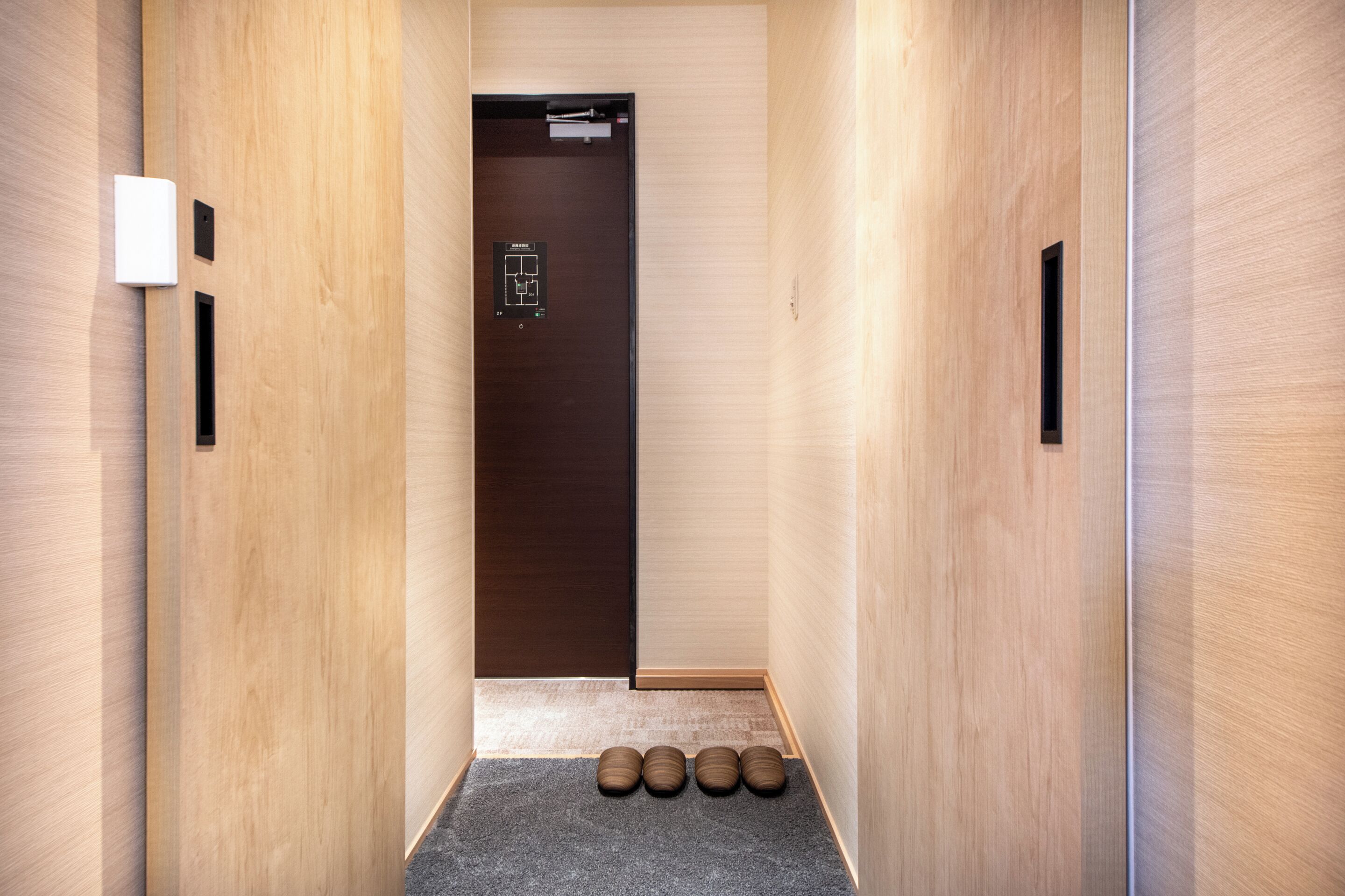 Un couloir étroit aux murs en bois, une porte en bois sombre au fond, et trois objets cylindriques de couleur marron posés sur un tapis gris.