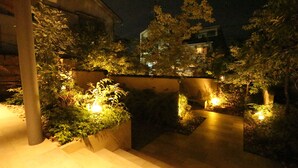 Un espace extérieur aménagé pour la nuit, avec des marches menant à un bâtiment, entouré de verdure et éclairé par un éclairage doux.