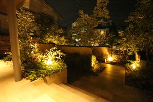 A nighttime outdoor area with steps leading up to a building, surrounded by greenery and illuminated by soft lighting.