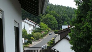 A balcony with a table and chairs, overlooking a residential street and a forested hill. - Tannenhof (Erlenbach am Main)