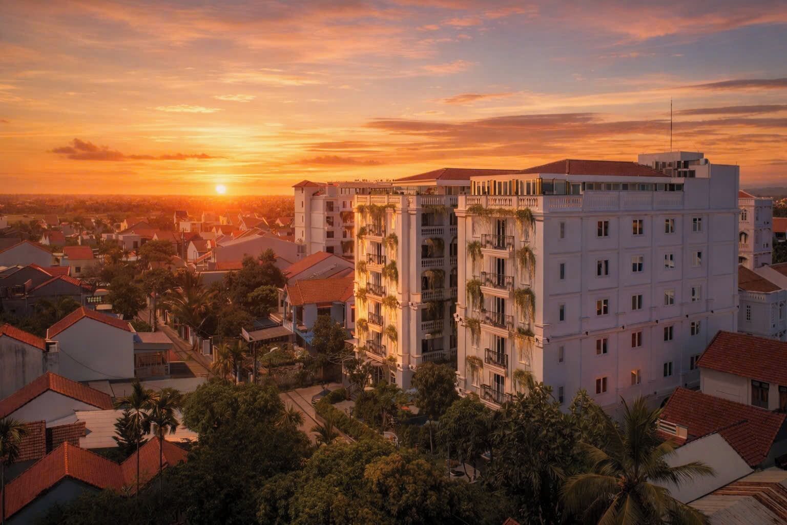 Un hotel de varios pisos con balcones, rodeado de edificios residenciales y palmeras, al atardecer.