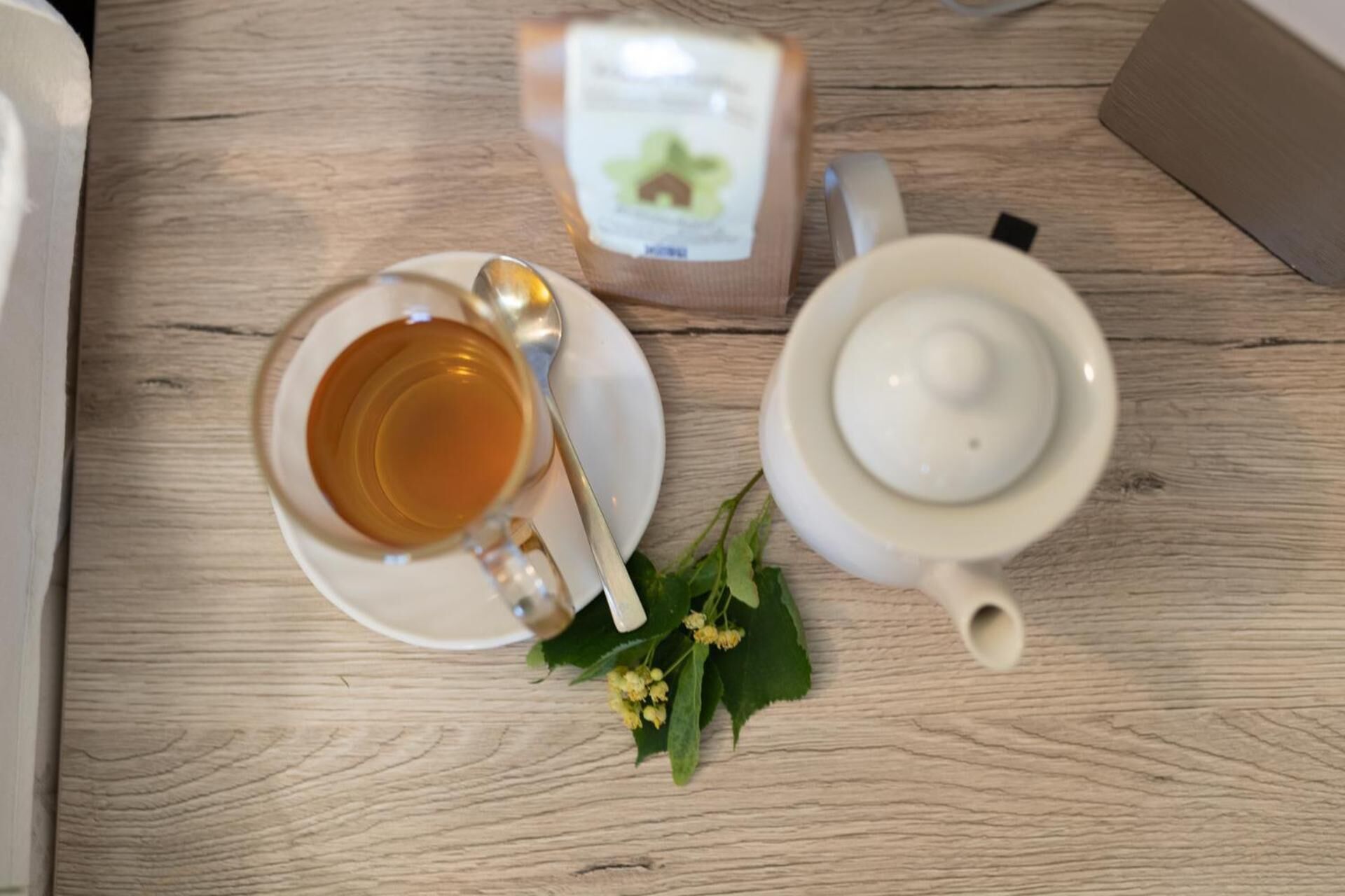 A wooden table with a white teapot, a cup of tea, and a spoon.