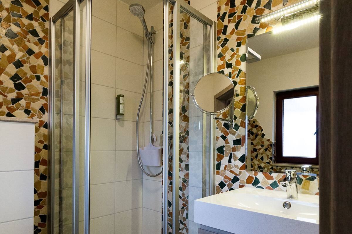 Un baño moderno con ducha acristalada, lavabo blanco con espejo y paredes con diseño de mosaicos.