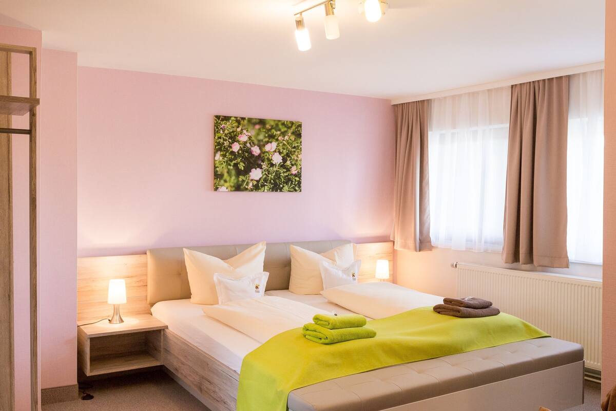 Una habitación de hotel con una cama, lámparas de noche, una toalla verde, una ventana con cortinas y un cuadro floral en la pared.
