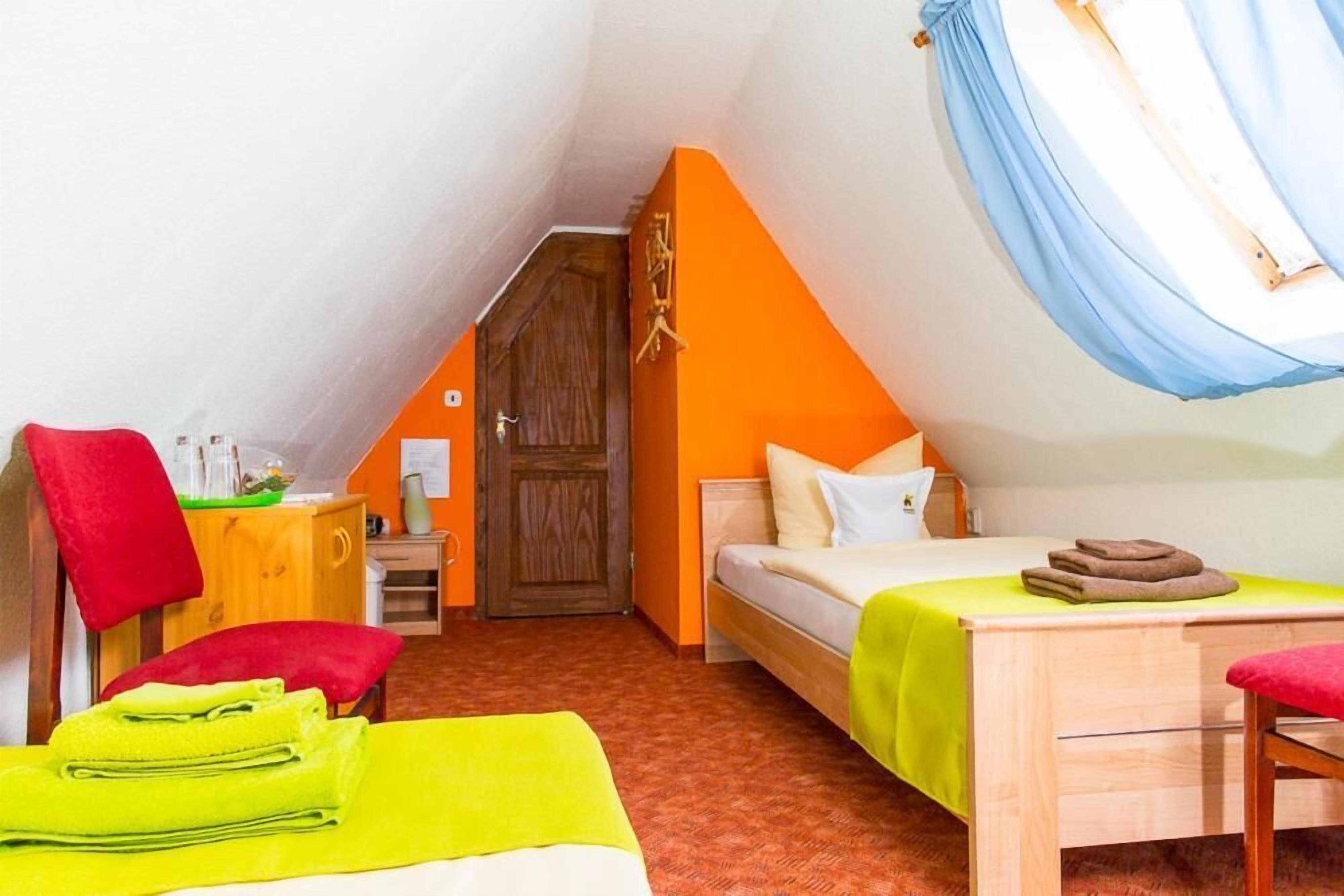 A small, cozy room with a sloped ceiling, a wooden door, a bed with a green bedspread, a small table, and a chair.