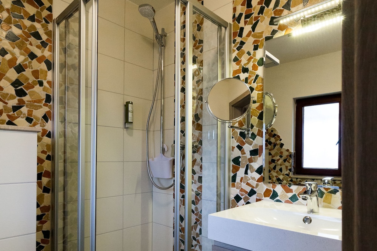 Un baño moderno con ducha acristalada, lavabo blanco con espejo y paredes con diseño de mosaicos.