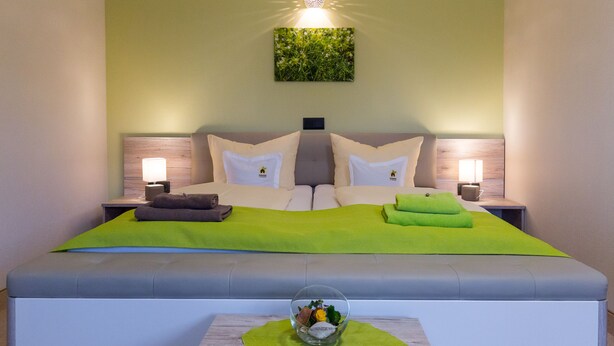 A modern hotel room with a large bed, green throw pillows, a bedside table with a lamp, and a wall-mounted light fixture. - Kräuterhotel Garni zur Klostergrotte (Grammetal)