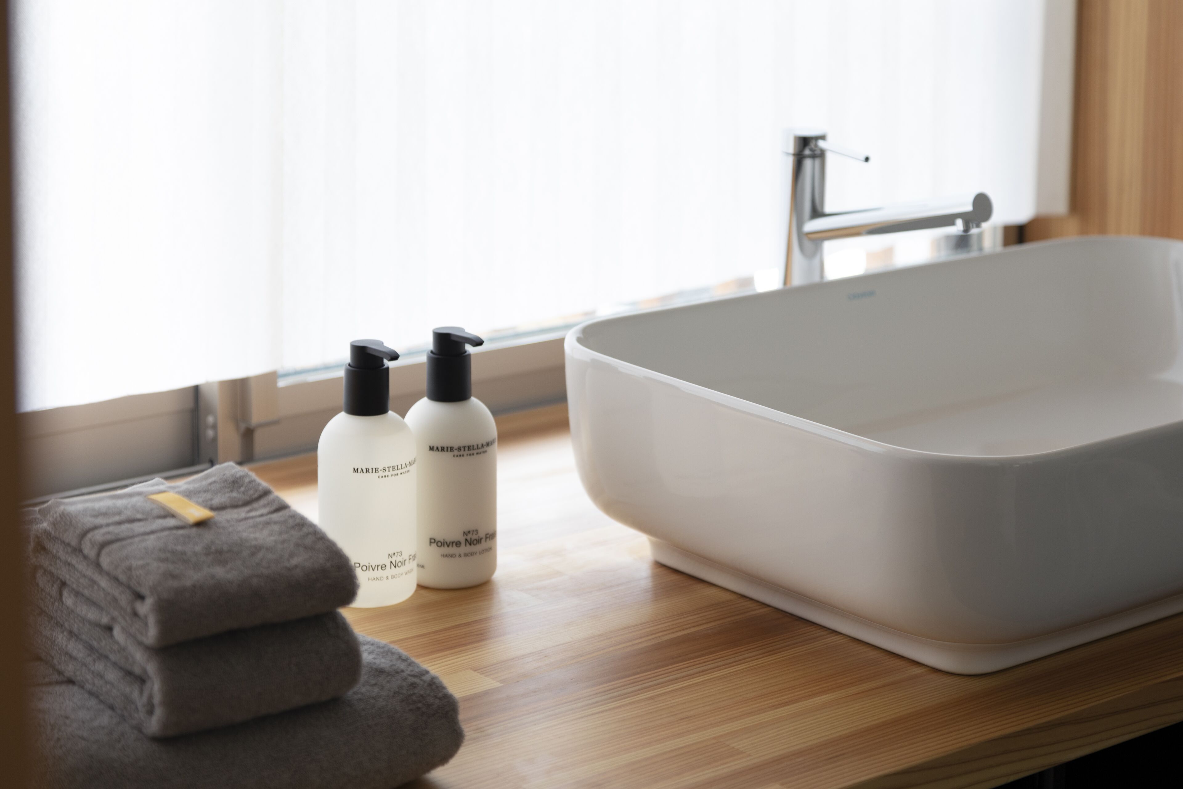 Un baño con un lavamanos blanco, dos frascos de productos para el cuidado de la piel y toallas dobladas sobre una encimera de madera.